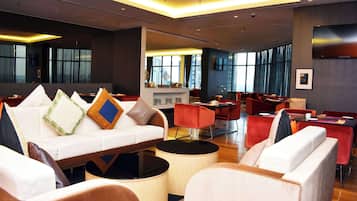 A modern hotel lobby with a sofa, red chairs, a bar area, and large windows.