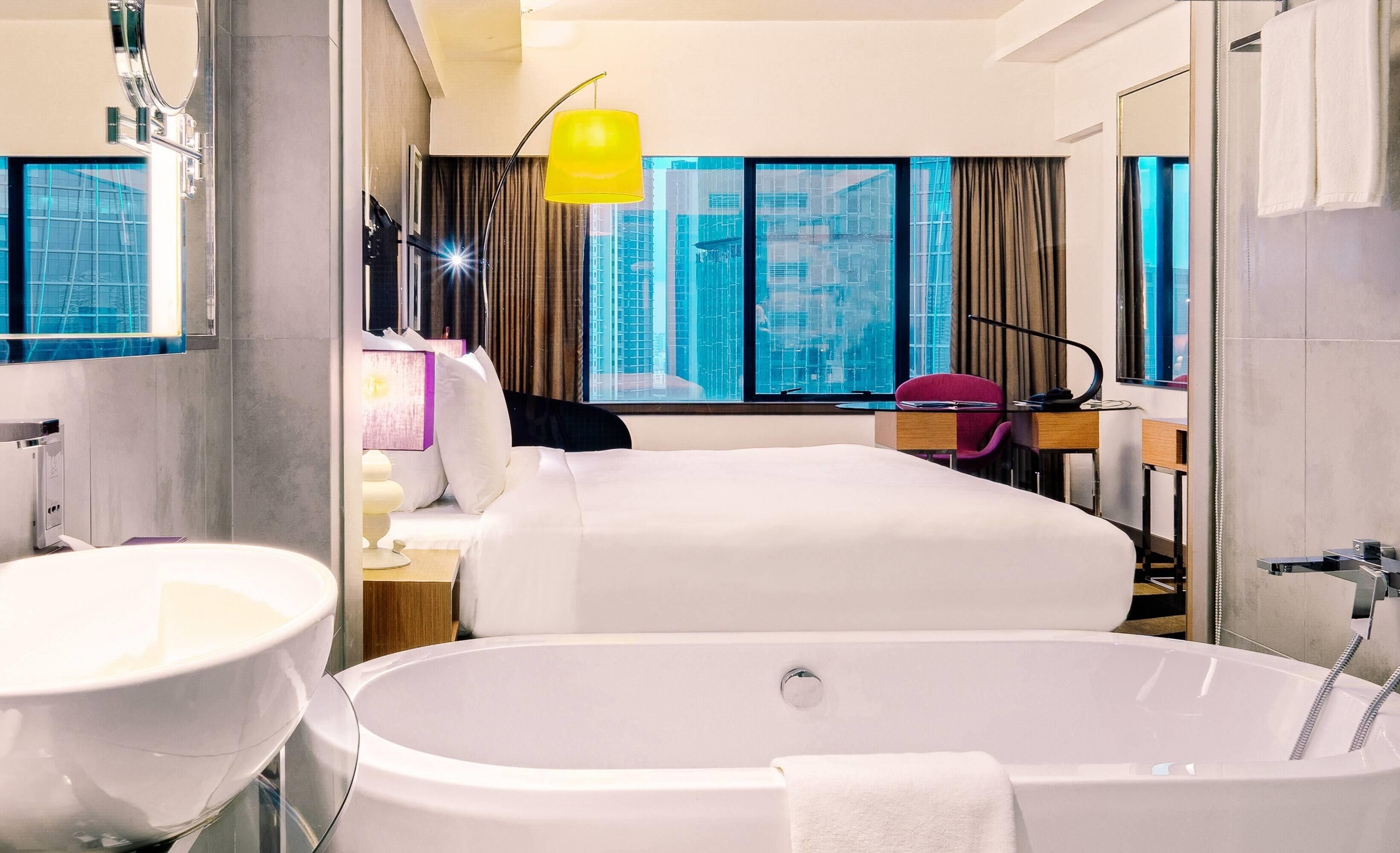 A hotel room with a large bathtub, a bed, a desk, and a view of tall buildings.