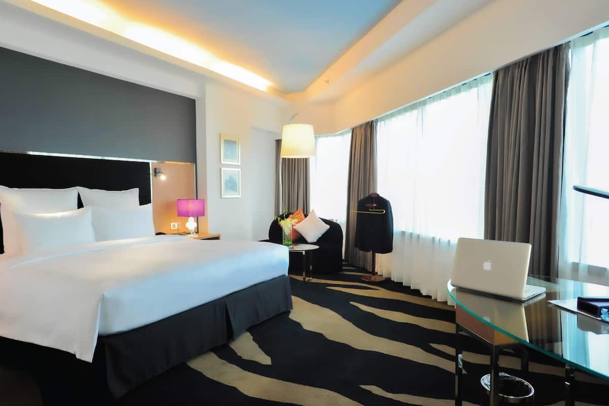 A hotel room with a bed, a glass table with a laptop, a chair, a lamp, a coat rack, and a window with curtains.