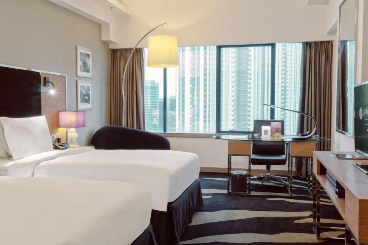 A hotel room with two beds, a desk, a chair, a TV, and a large window with a city view.
