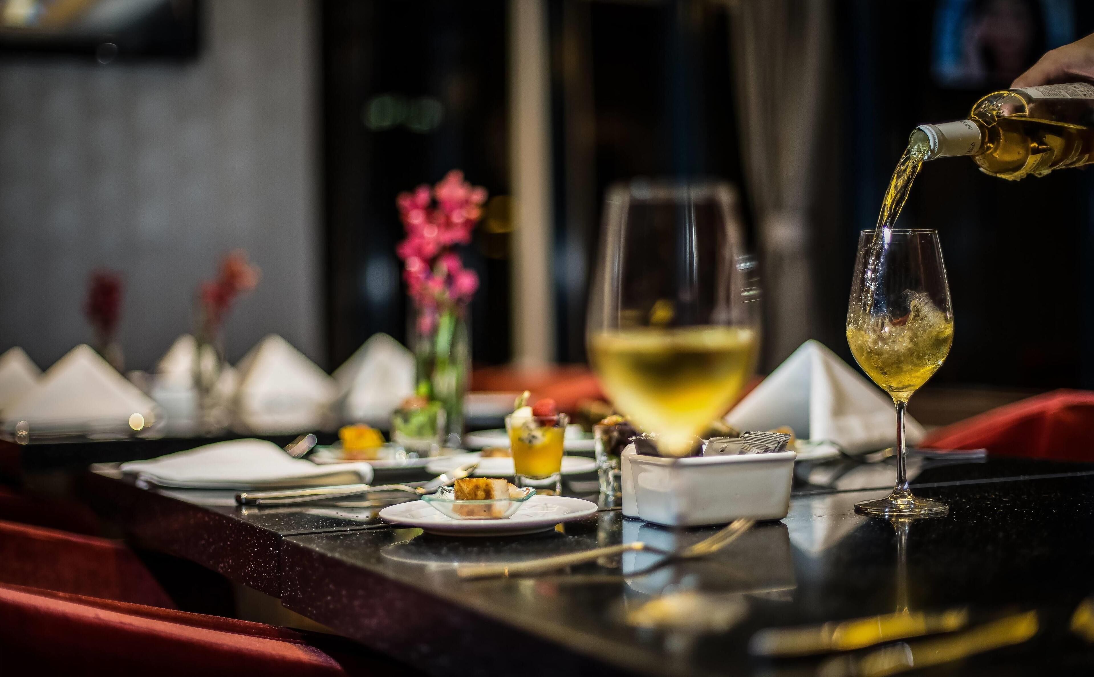A dining table with a glass of white wine being poured, surrounded by various dishes and a vase of flowers.