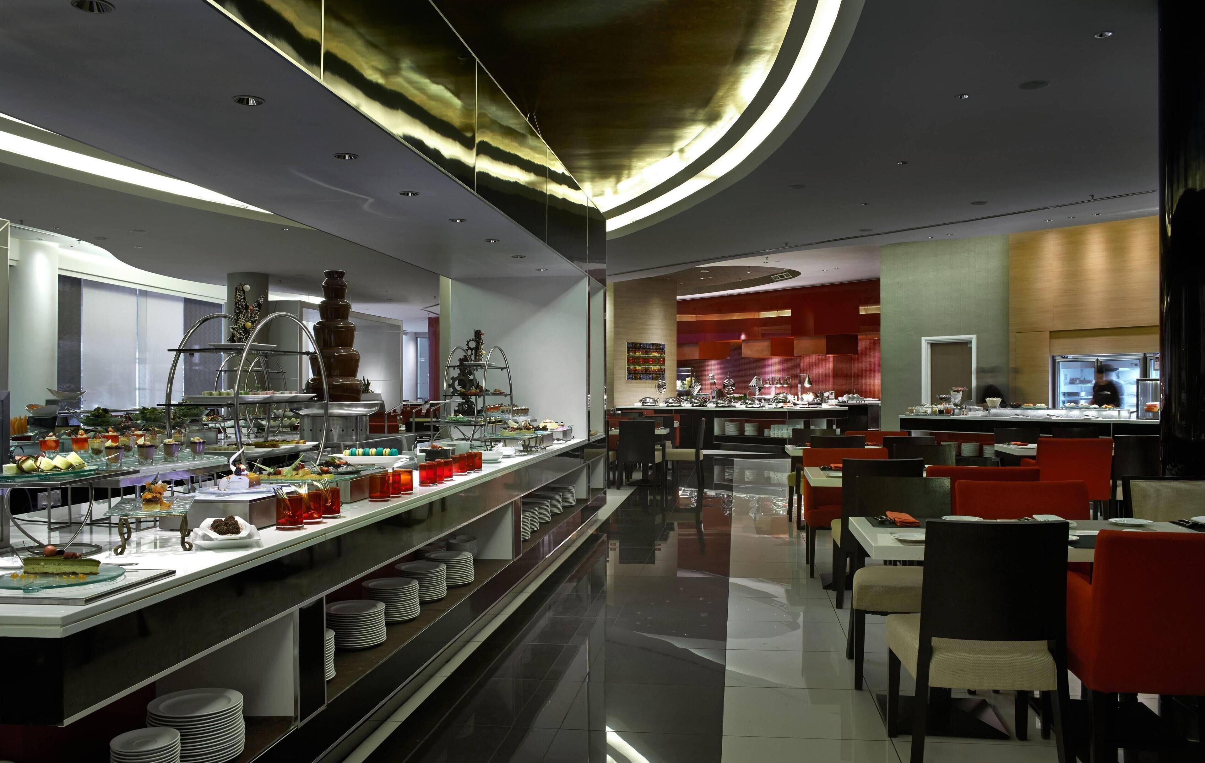 A modern restaurant with a buffet setup, multiple dining tables, and a kitchen area in the background.