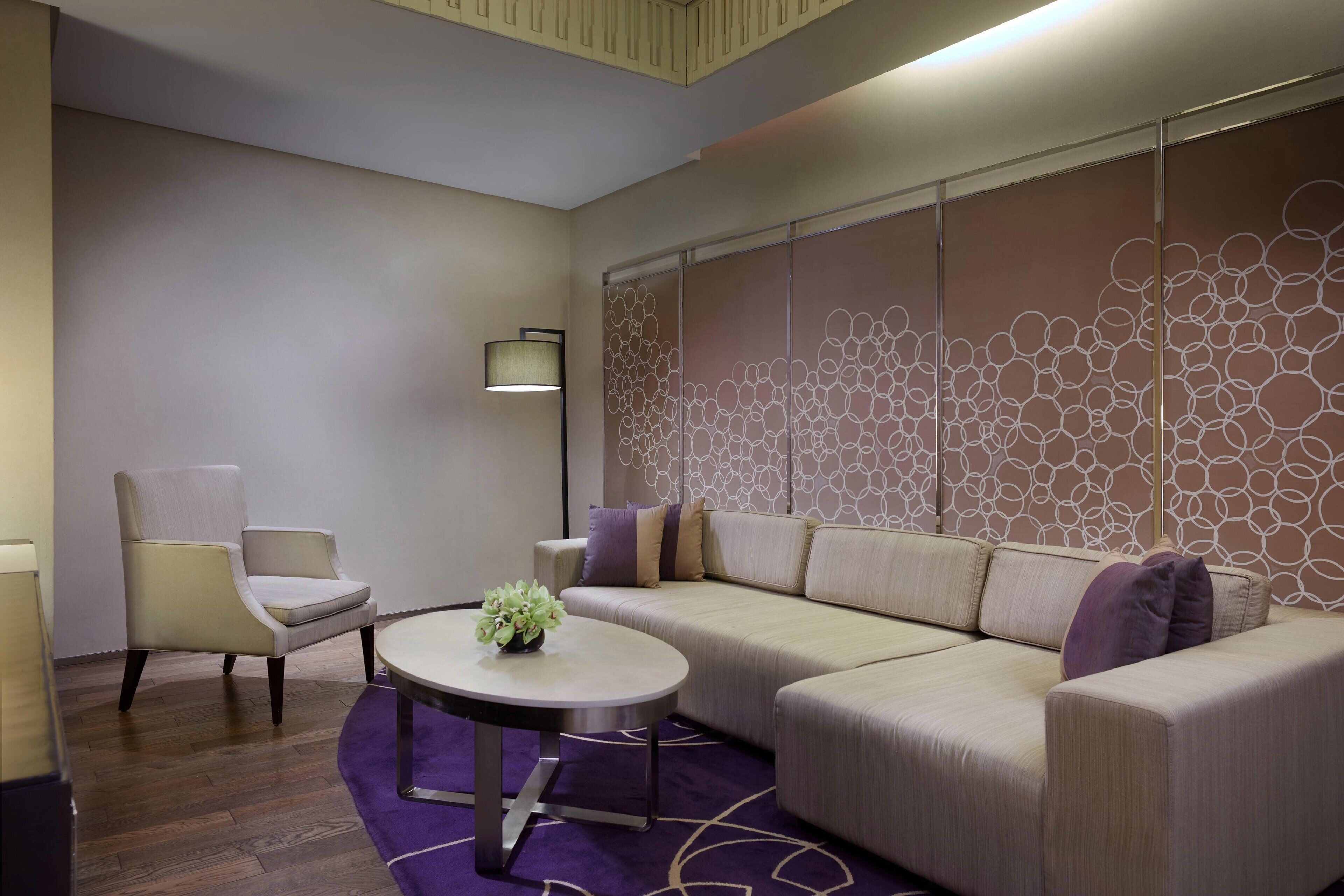 Una habitación de hotel moderna con un sofá color beige, una mesa redonda con una planta, una silla y un panel decorativo en la pared.