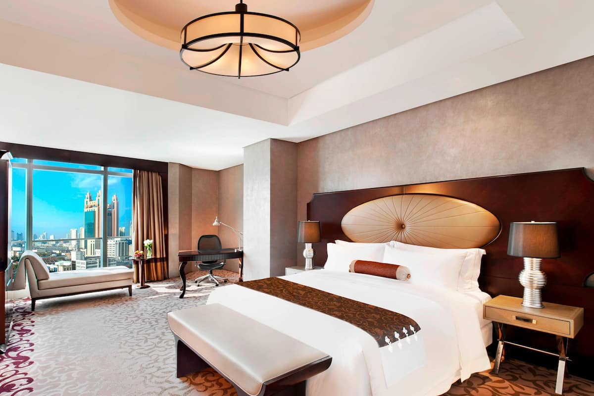 Ein Hotelzimmer mit einem großen Bett, einem Schreibtisch, einem Sessel, einer Bank, einem Nachttisch und Blick auf die Stadt.