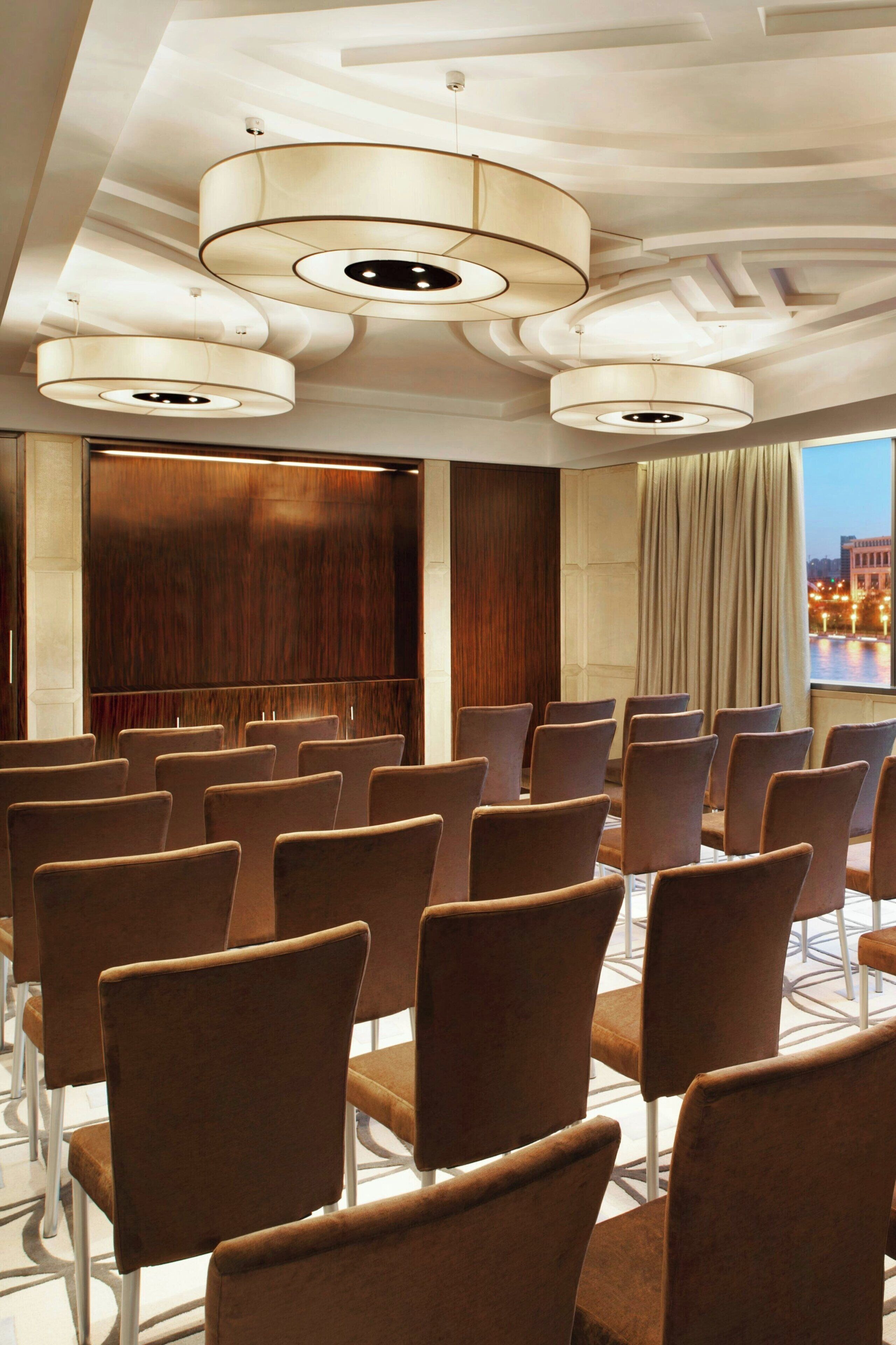 Un salón de conferencias con filas de sillas marrones, un panel de pared de madera y un amplio ventanal con vistas a un paisaje urbano.