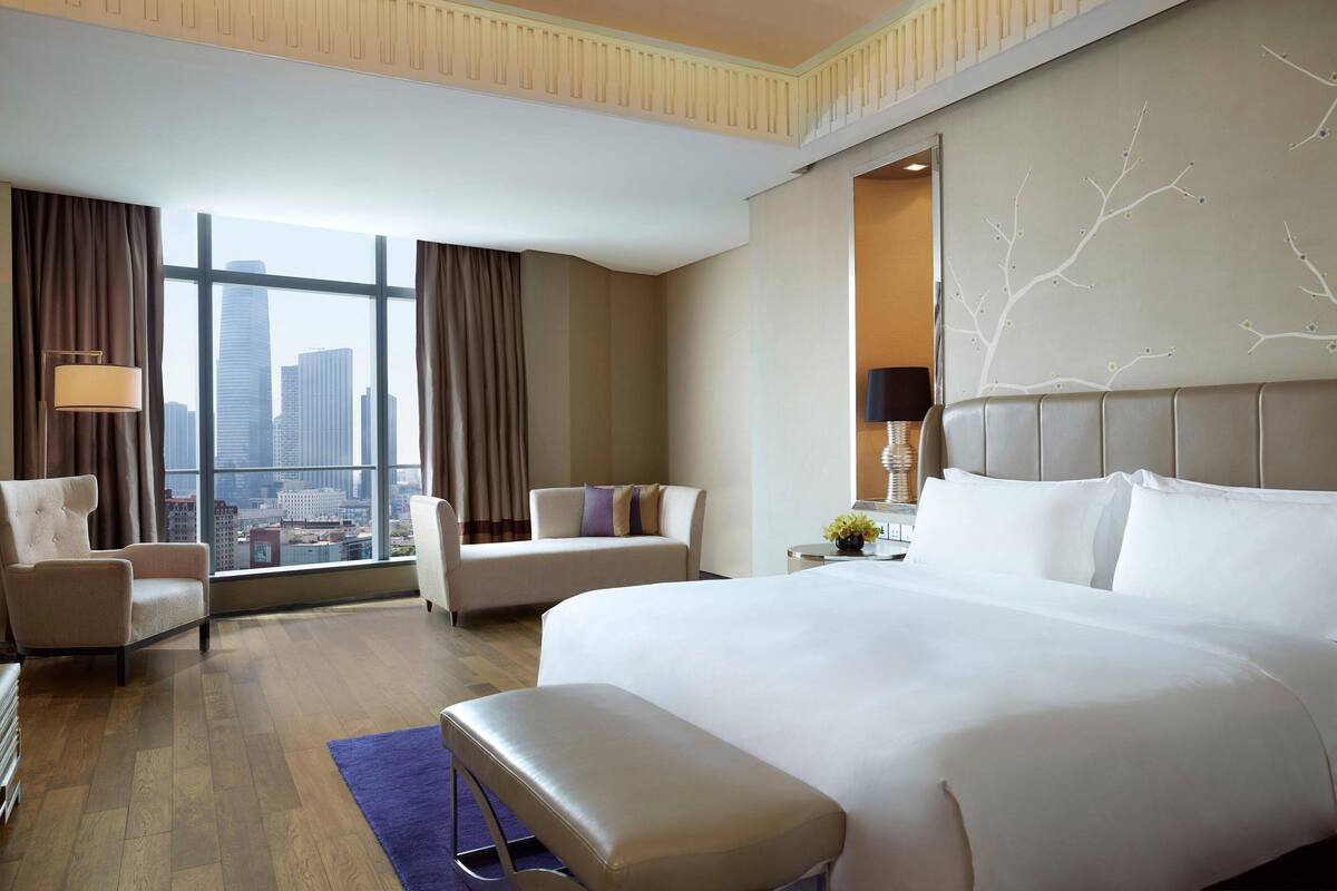 Ein Hotelzimmer mit einem großen Bett, zwei Sesseln, einer Bank und Blick auf die Stadt durch große Fenster.