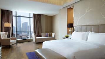 Ein Hotelzimmer mit einem großen Bett, zwei Sesseln, einer Bank und Blick auf die Stadt durch große Fenster.