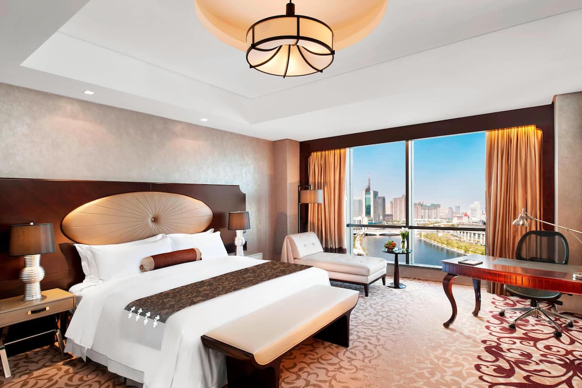 Ein Hotelzimmer mit einem großen Bett, einem Schreibtisch, einem Stuhl und Blick auf eine Stadtlandschaft durch große Fenster.