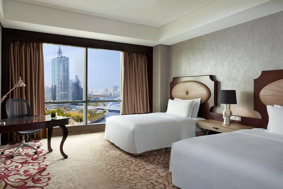 Ein Hotelzimmer mit zwei Betten, einem Schreibtisch und Blick auf eine Stadtlandschaft.