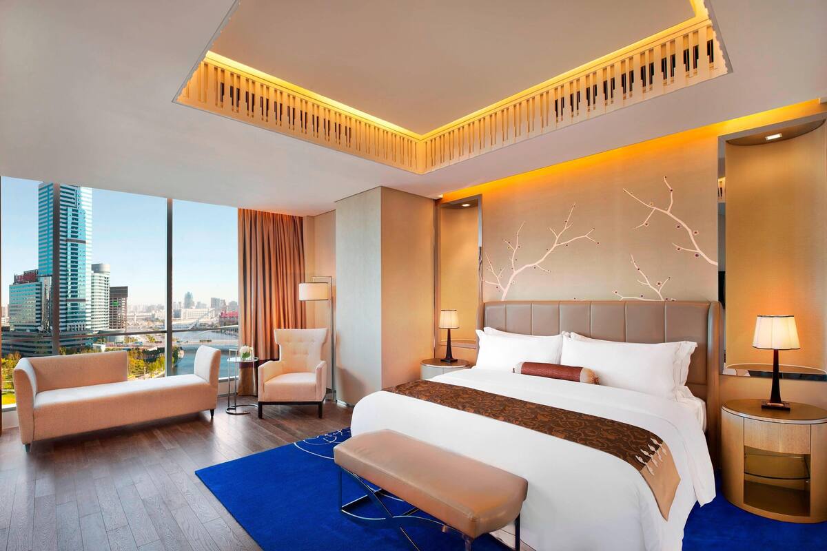 Ein modernes Hotelzimmer mit einem großen Bett, einer Couch, einem Sessel und Blick auf die Skyline.