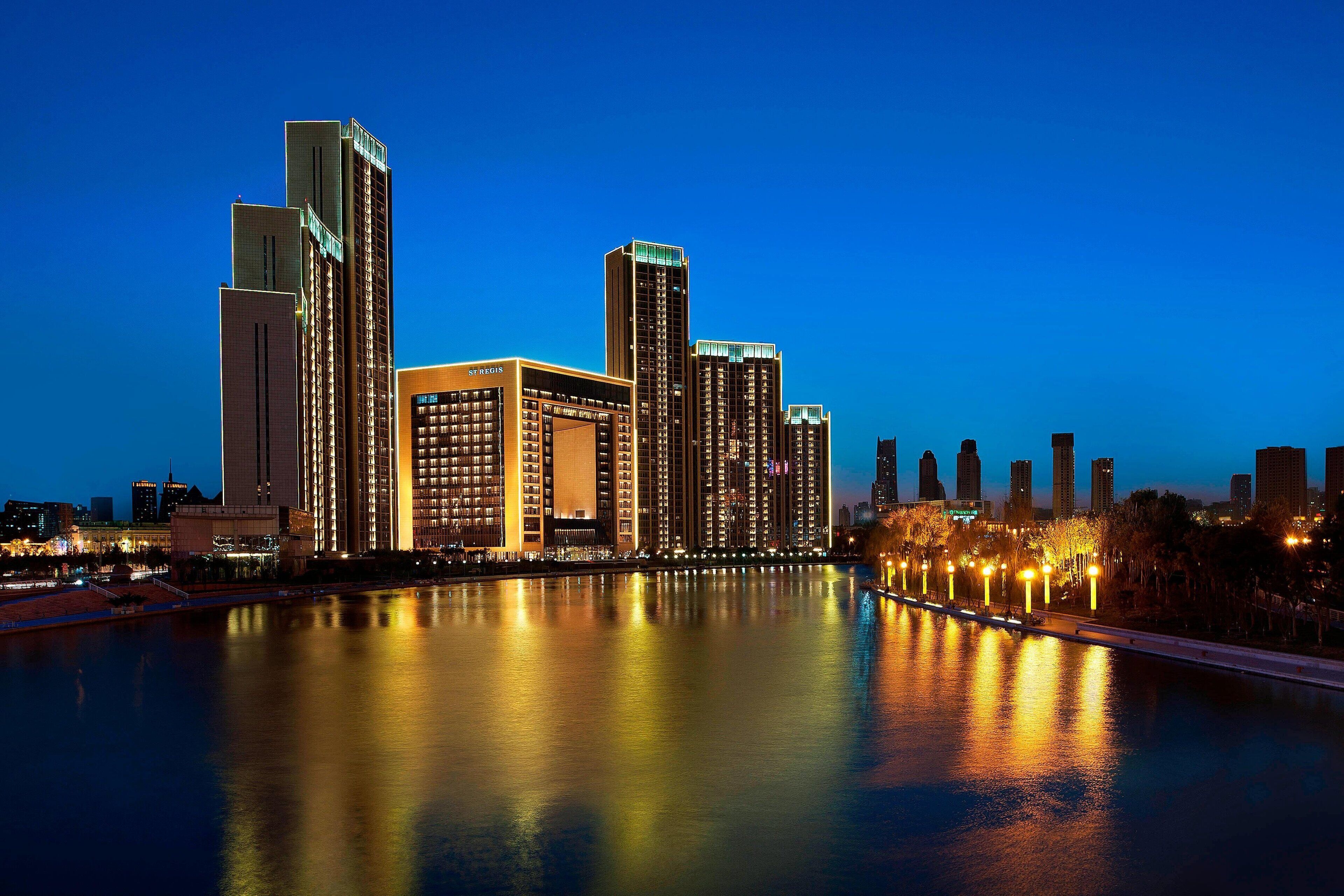 A high-rise hotel complex with multiple buildings along a waterfront, featuring illuminated windows and a reflection in the water.