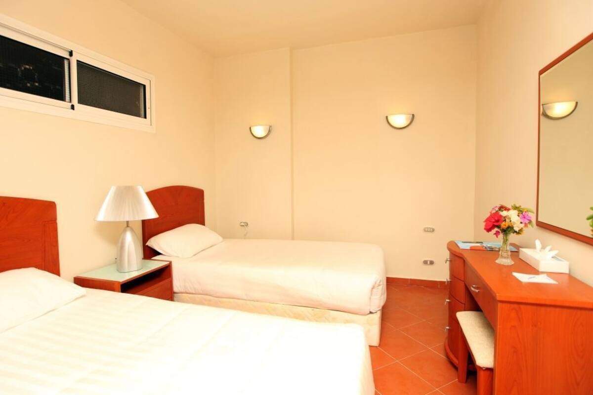 Une chambre d’hôtel avec deux lits, une table de chevet avec une lampe, une commode en bois, un miroir et une fenêtre.