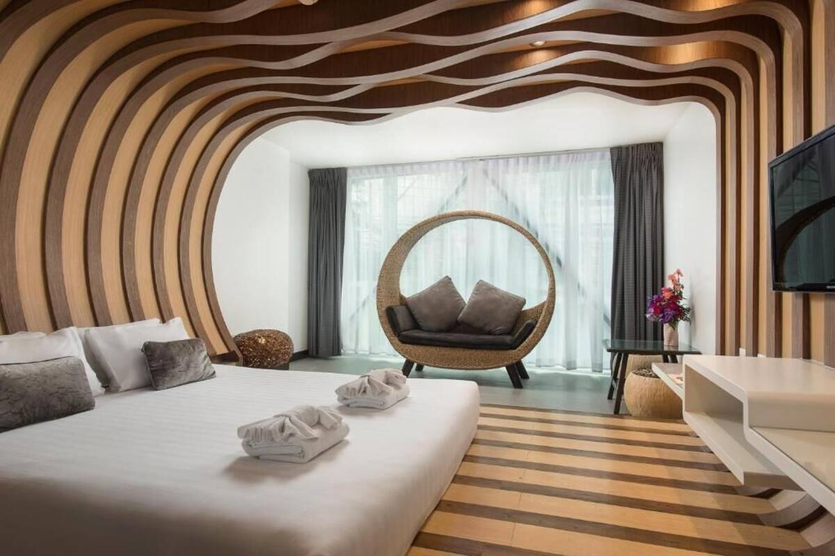 Une chambre d’hôtel moderne dotée d’un grand lit, d’un fauteuil rond en osier, d’un téléviseur à écran plat et offrant une vue sur l’extérieur grâce à de grandes fenêtres.