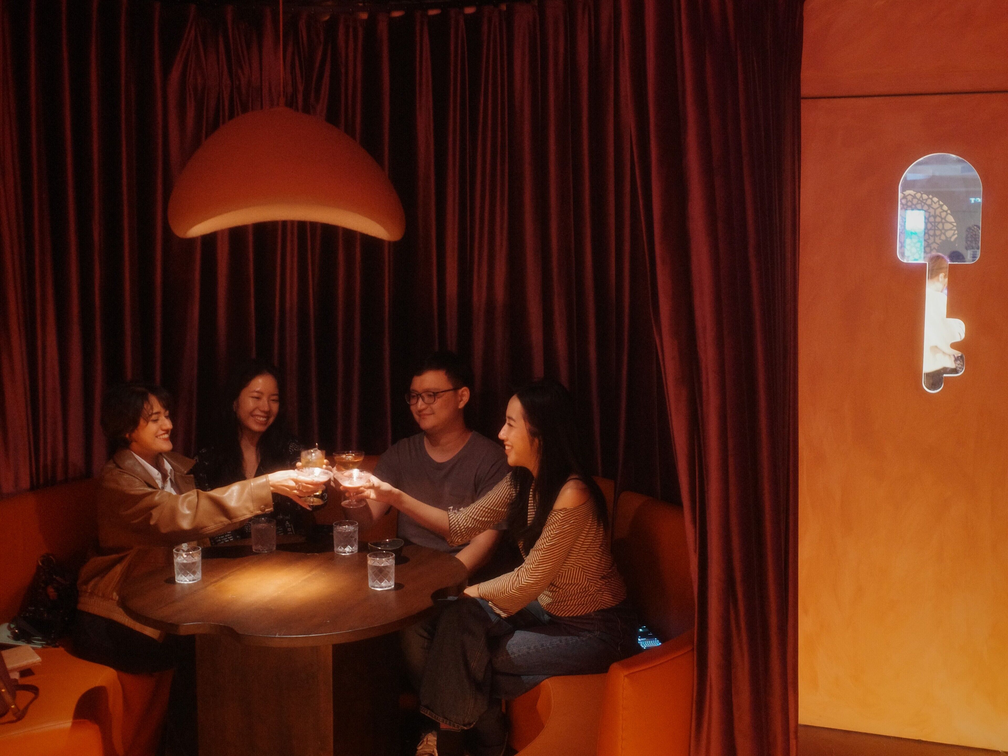 A group of people seated around a round table in a dimly lit room with red curtains and a large pendant light.