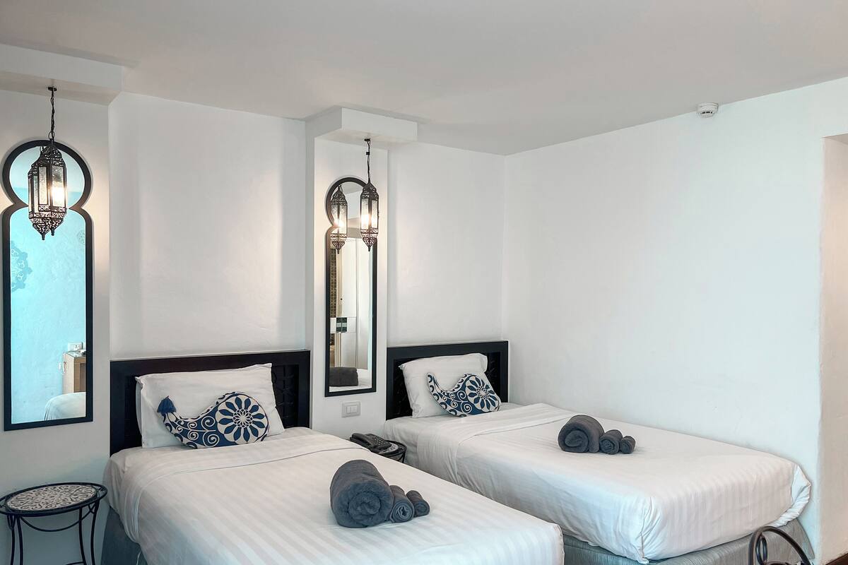Une chambre d’hôtel avec deux lits, chacun recouvert d’une courtepointe blanche et agrémenté d’oreillers assortis, une petite table ronde et un miroir avec un cadre décoratif.