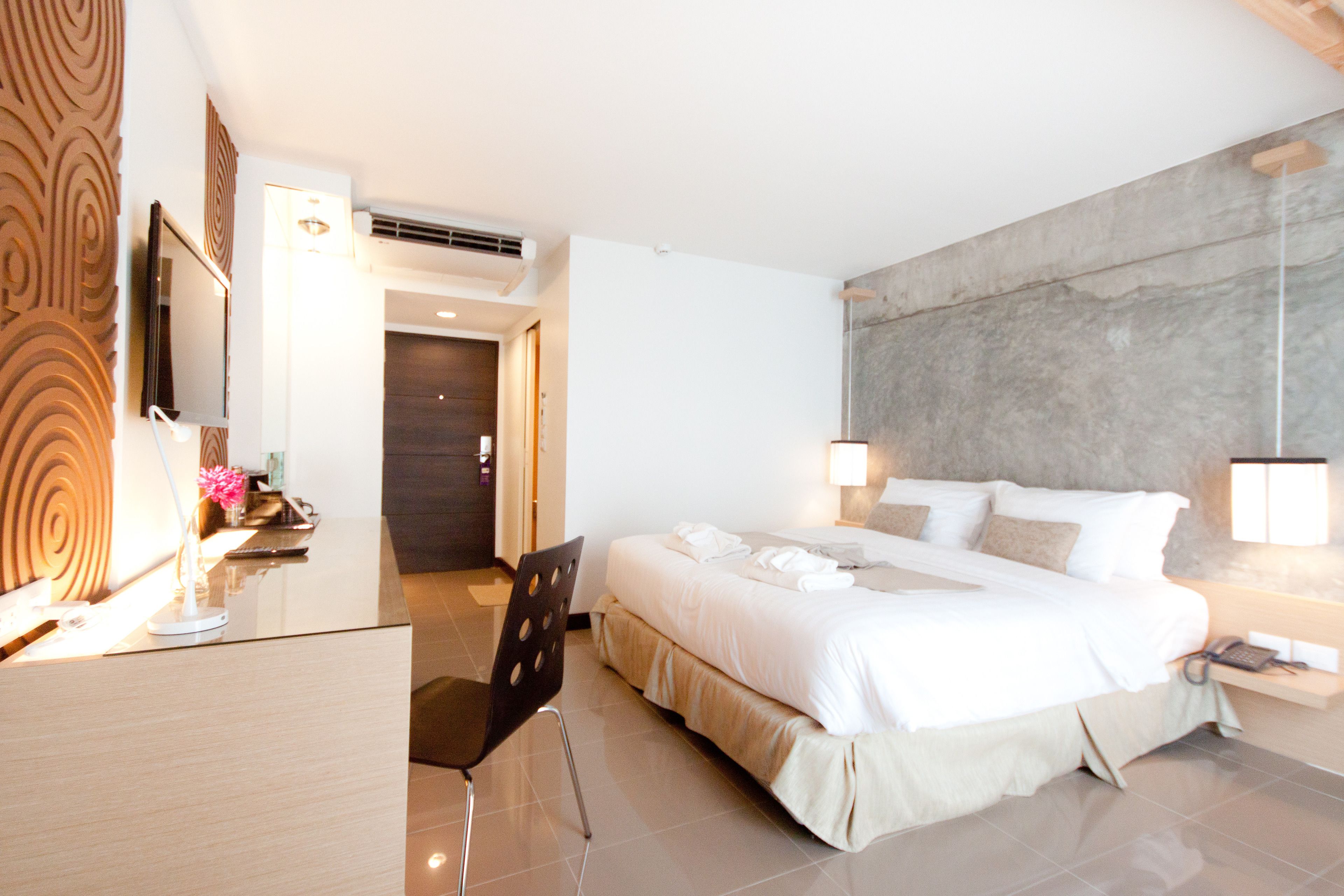 A modern hotel room with a large bed, a desk, and a chair. There is a mirror on the wall and a lamp on the desk.