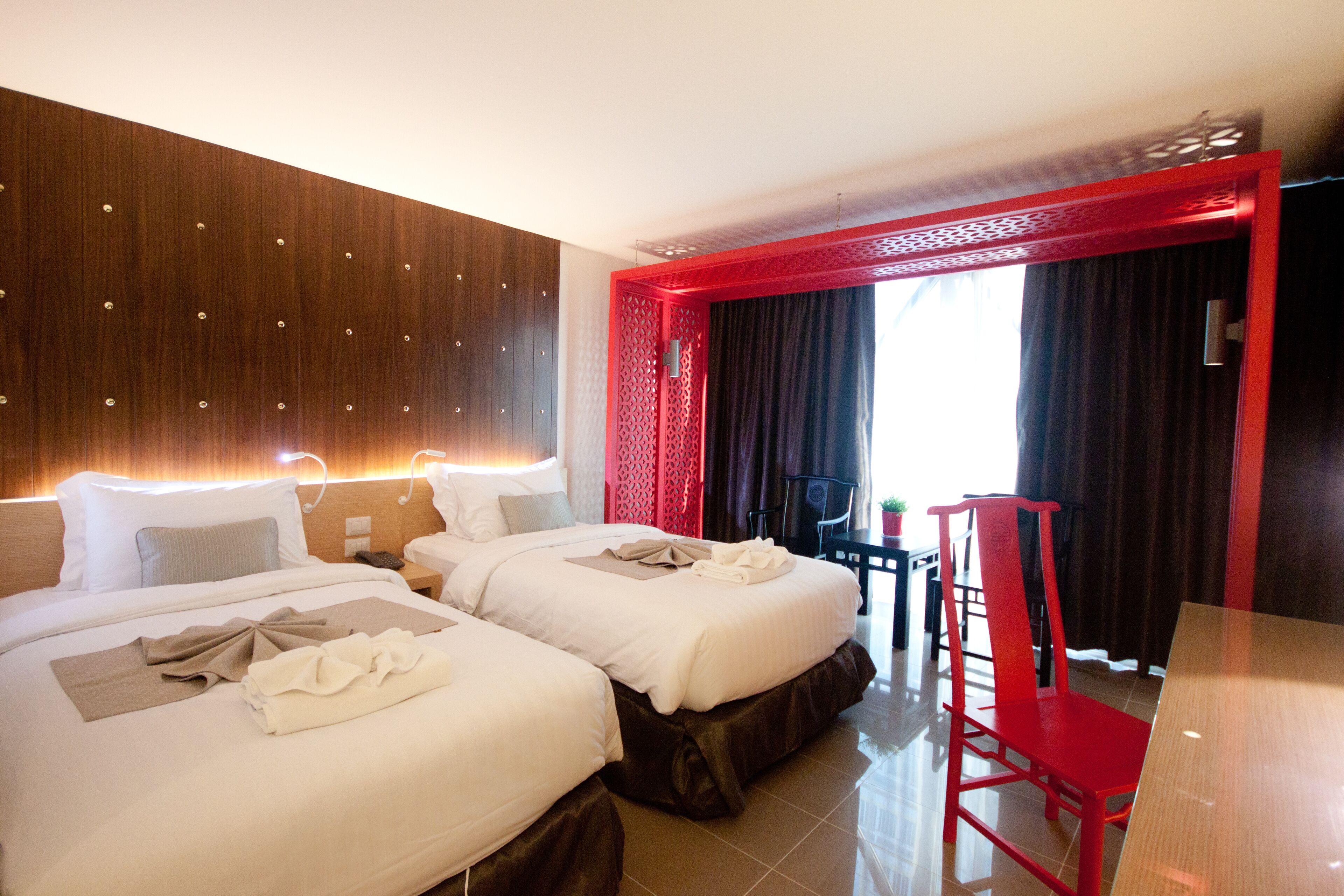 A hotel room with two beds, a red accent wall, a small table, and a chair.
