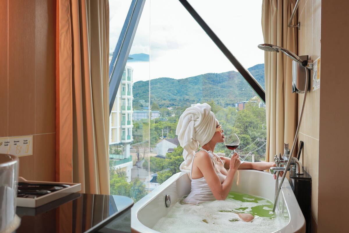 Une personne dans une baignoire, une serviette sur la tête, tenant un verre de vin, et un pommeau de douche au-dessus de la baignoire.