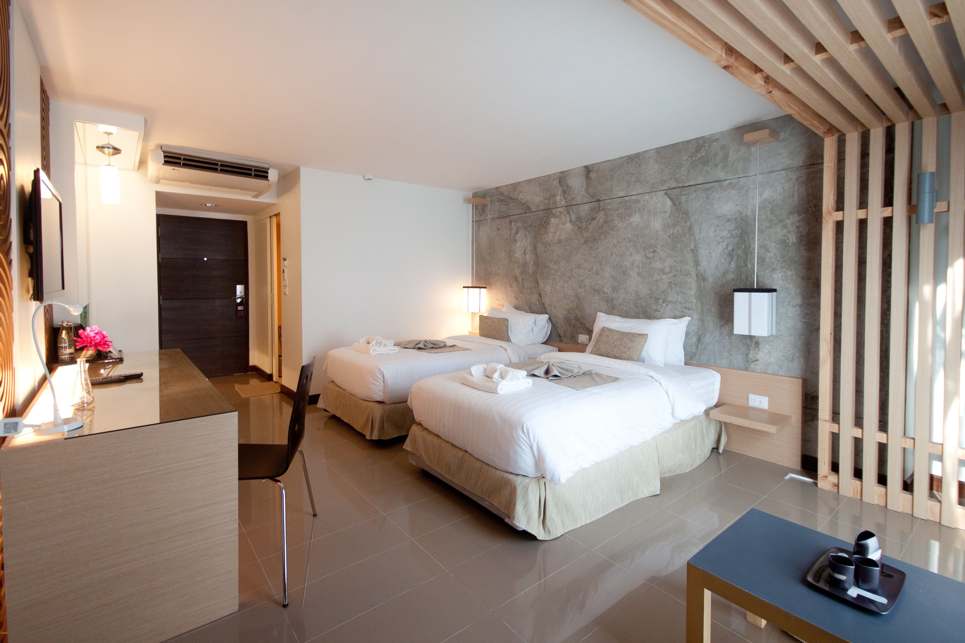 A hotel room with two beds, a desk, and a chair. There is a desk lamp, a small table with a tray, and a wall-mounted air conditioner.
