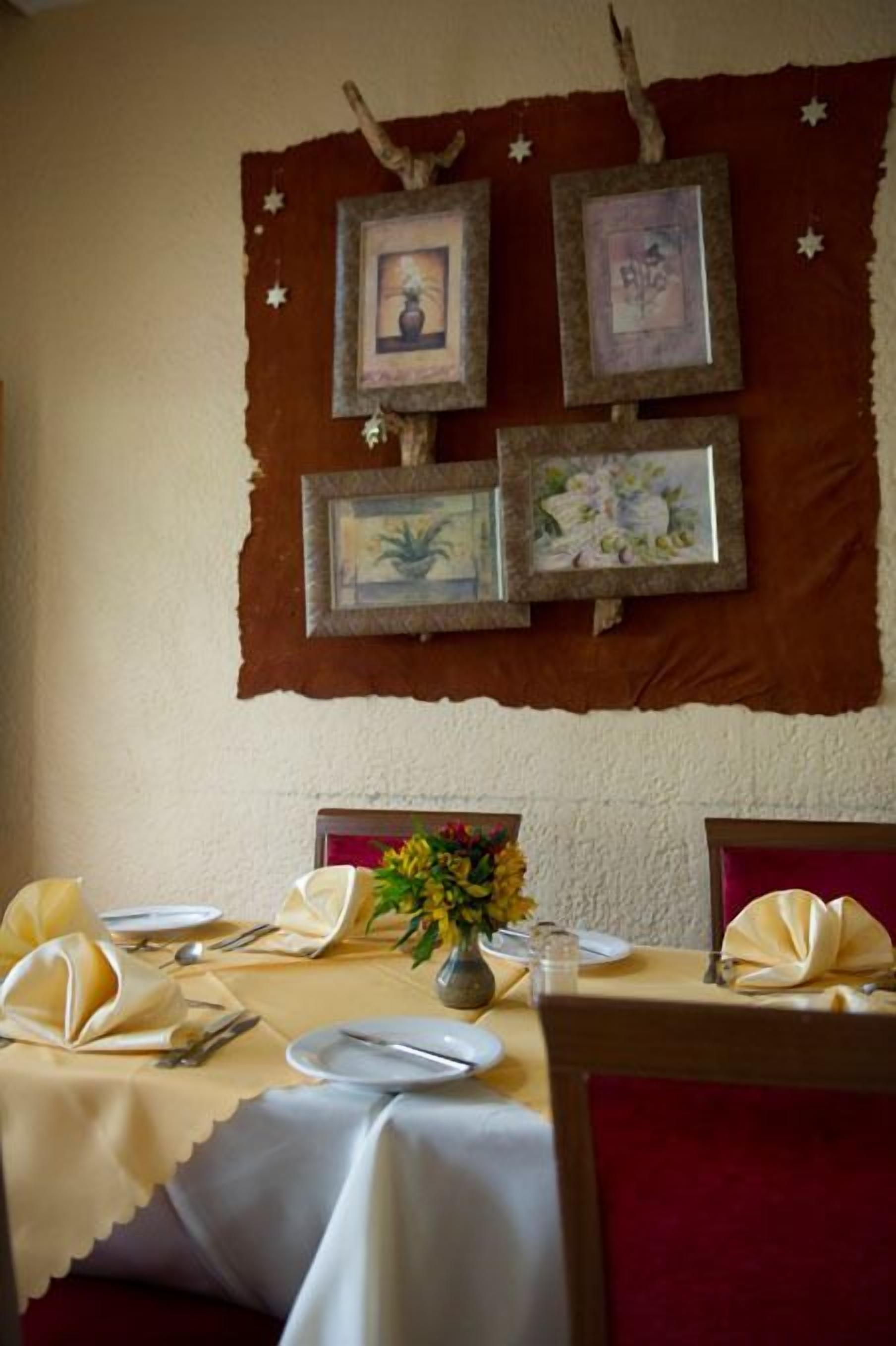 A dining area with a table set for two, framed pictures on the wall, and a vase with flowers on the table.