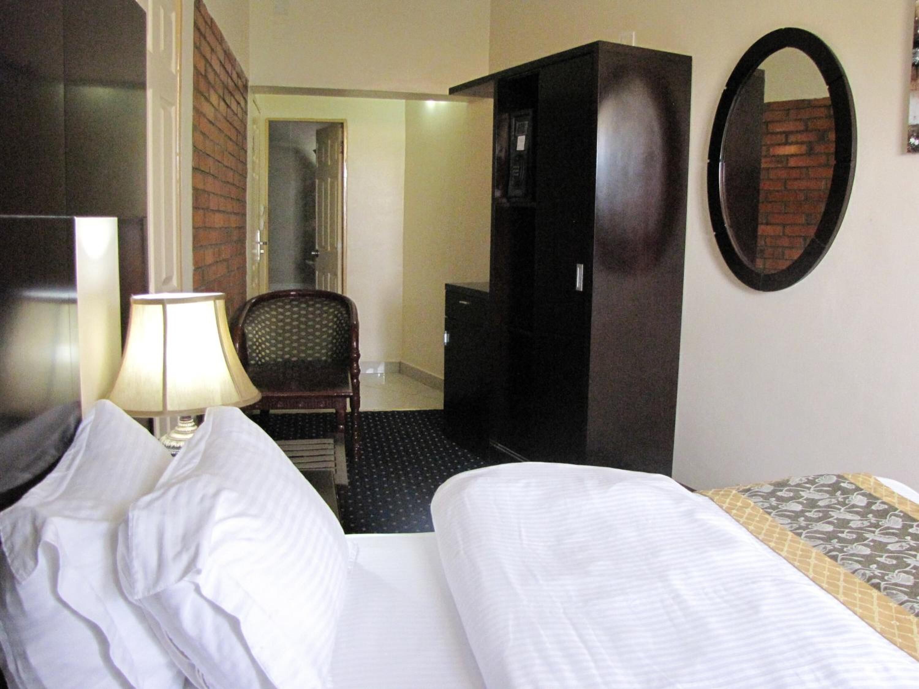 A hotel room with a bed, a chair, a desk, a lamp, a mirror, and a wardrobe.