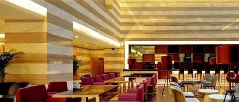 A spacious lobby with a long reception desk, multiple seating areas with red chairs, and a modern design with wooden flooring and striped walls.