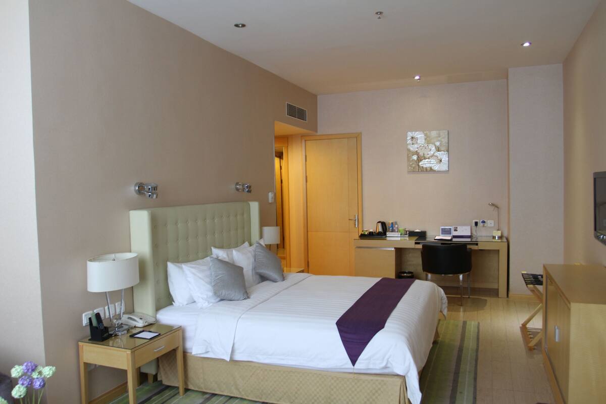 A hotel room with a bed, bedside table with a lamp, a desk with a computer, and a television.