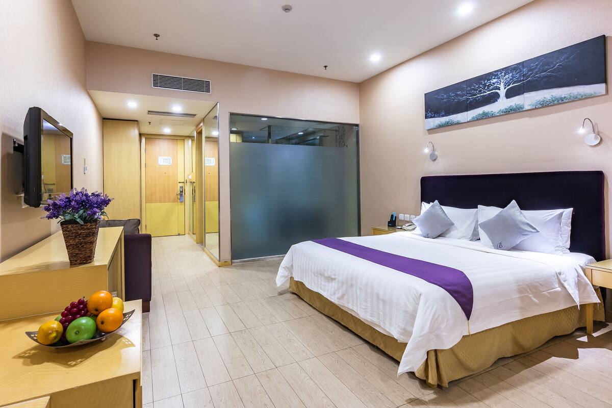 A hotel room with a bed, a desk with a fruit bowl, a mirror, and a painting on the wall.
