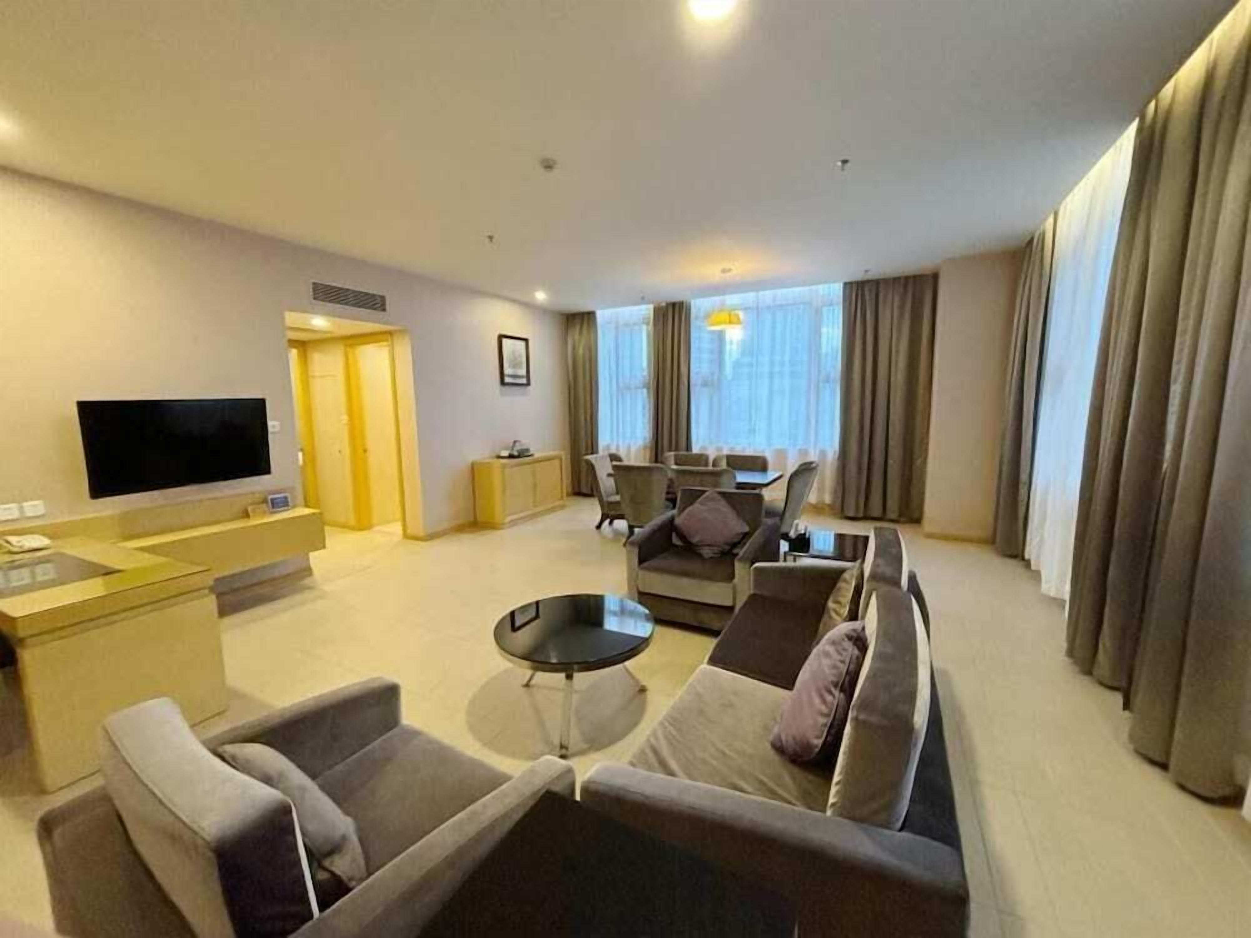 A modern hotel room with a living area featuring a sofa set, a coffee table, and a flat-screen TV. There is a small dining table and chairs in the background.
