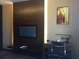 A hotel room with a flat-screen TV mounted on a wooden wall, a desk with a chair, and a framed abstract painting.