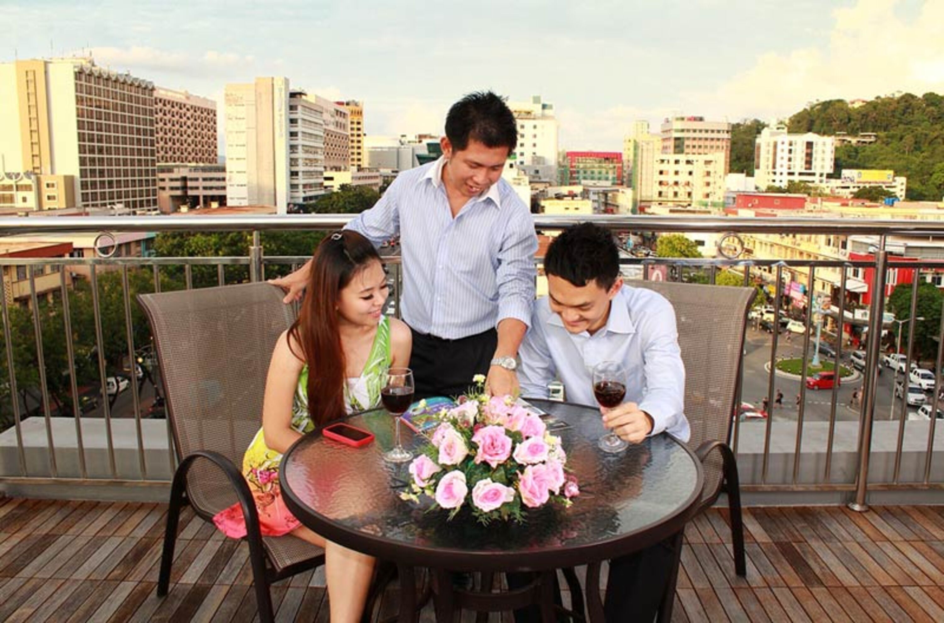 A balcony with a table set for three people, a floral centerpiece, and a cityscape view.