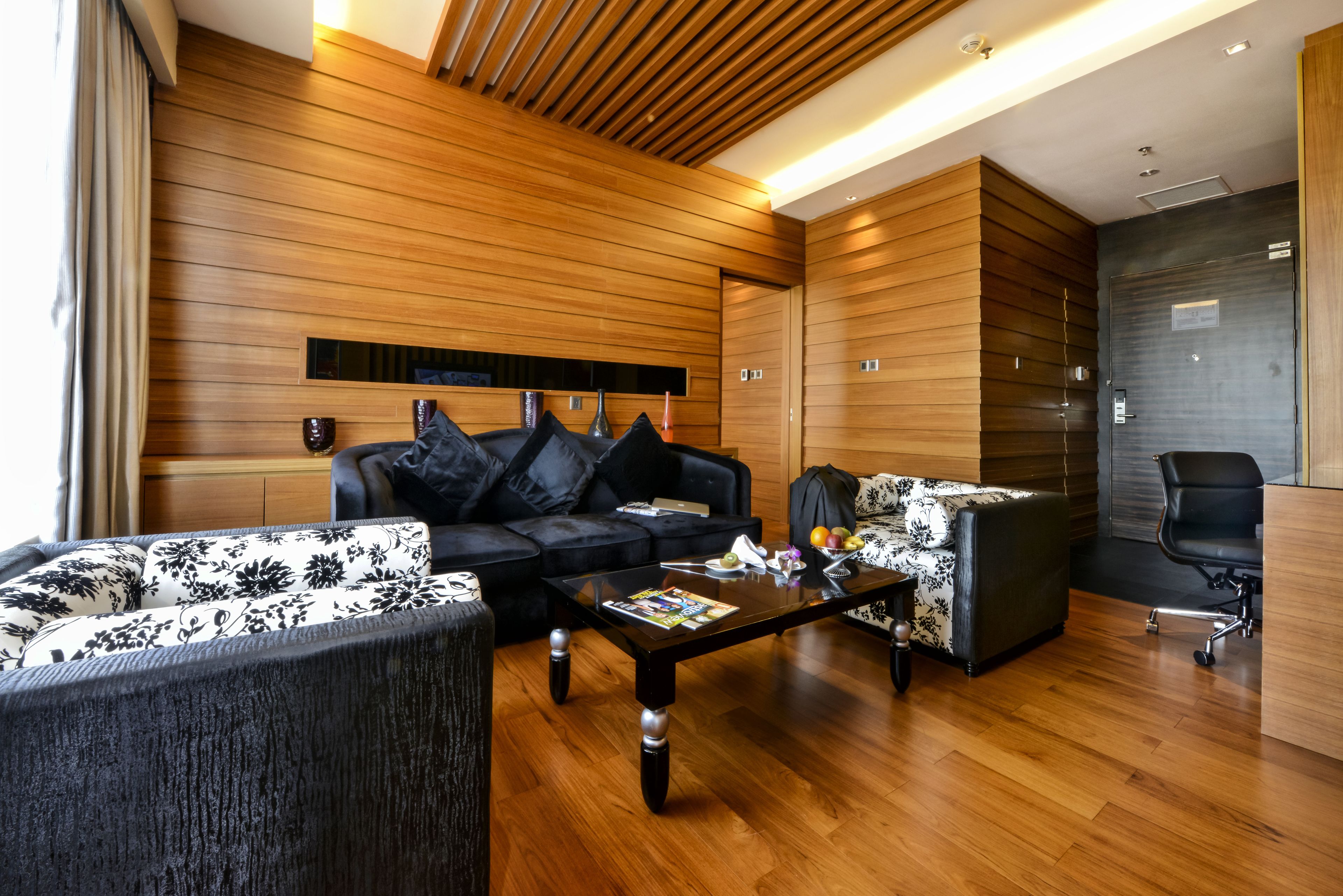A modern living room with wooden panel walls, a black sofa, a floral-patterned sofa, and a glass coffee table.