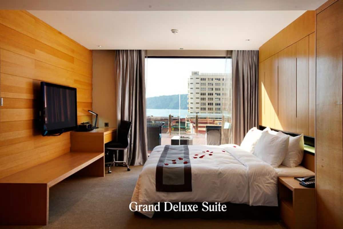 Eine Grand Deluxe Suite mit Bett, Wand-Fernseher, Schreibtisch und Blick auf die Stadt.