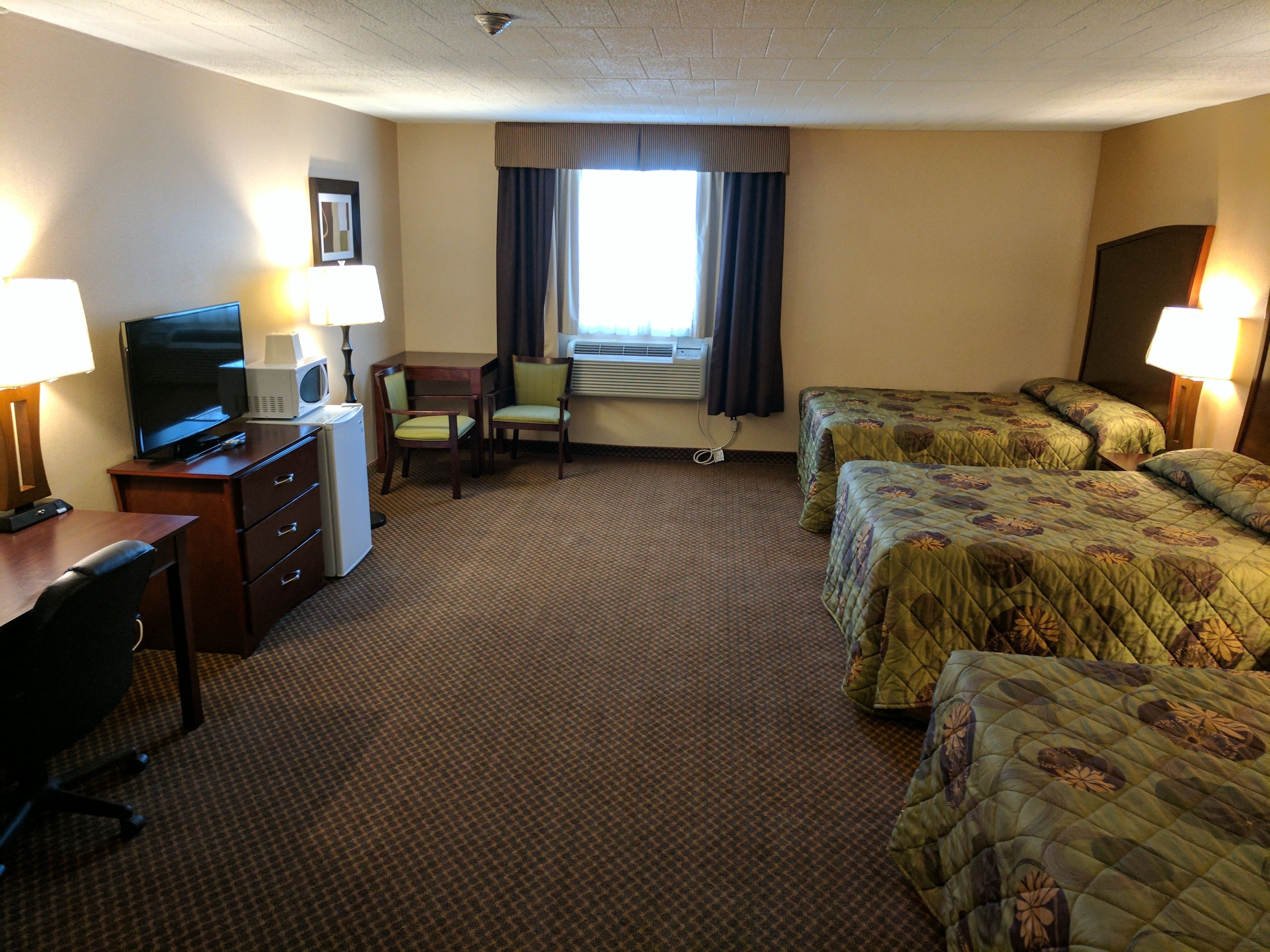 A hotel room with a television, desk, two chairs, a desk lamp, two beds with floral bedding, an air conditioning unit, and a window with curtains.