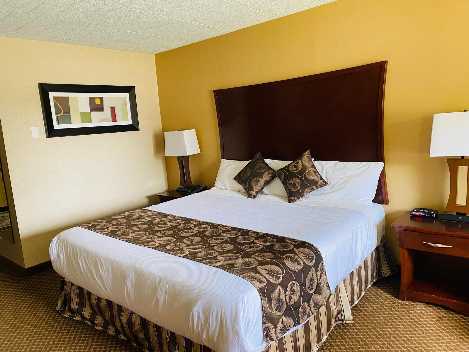 A hotel room with a large bed, two bedside tables, a nightstand, a framed picture on the wall, and a carpeted floor.