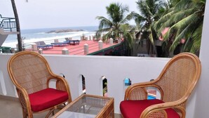 A balcony with wicker chairs and a glass table, overlooking a beach with palm trees and a red-roofed structure. - Jeevan Ayurvedic Beach Resort (Thiruvananthapuram)
