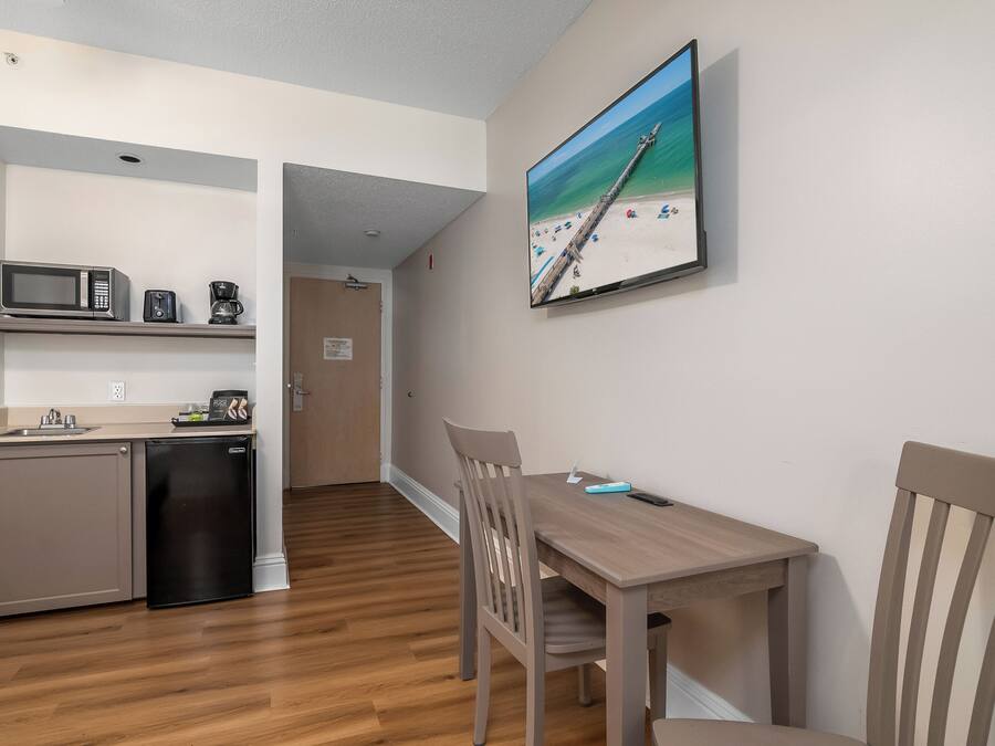 A compact kitchen with a microwave, toaster, and coffee maker, a dining area with a table and chairs, and a beach-themed TV on the wall.
