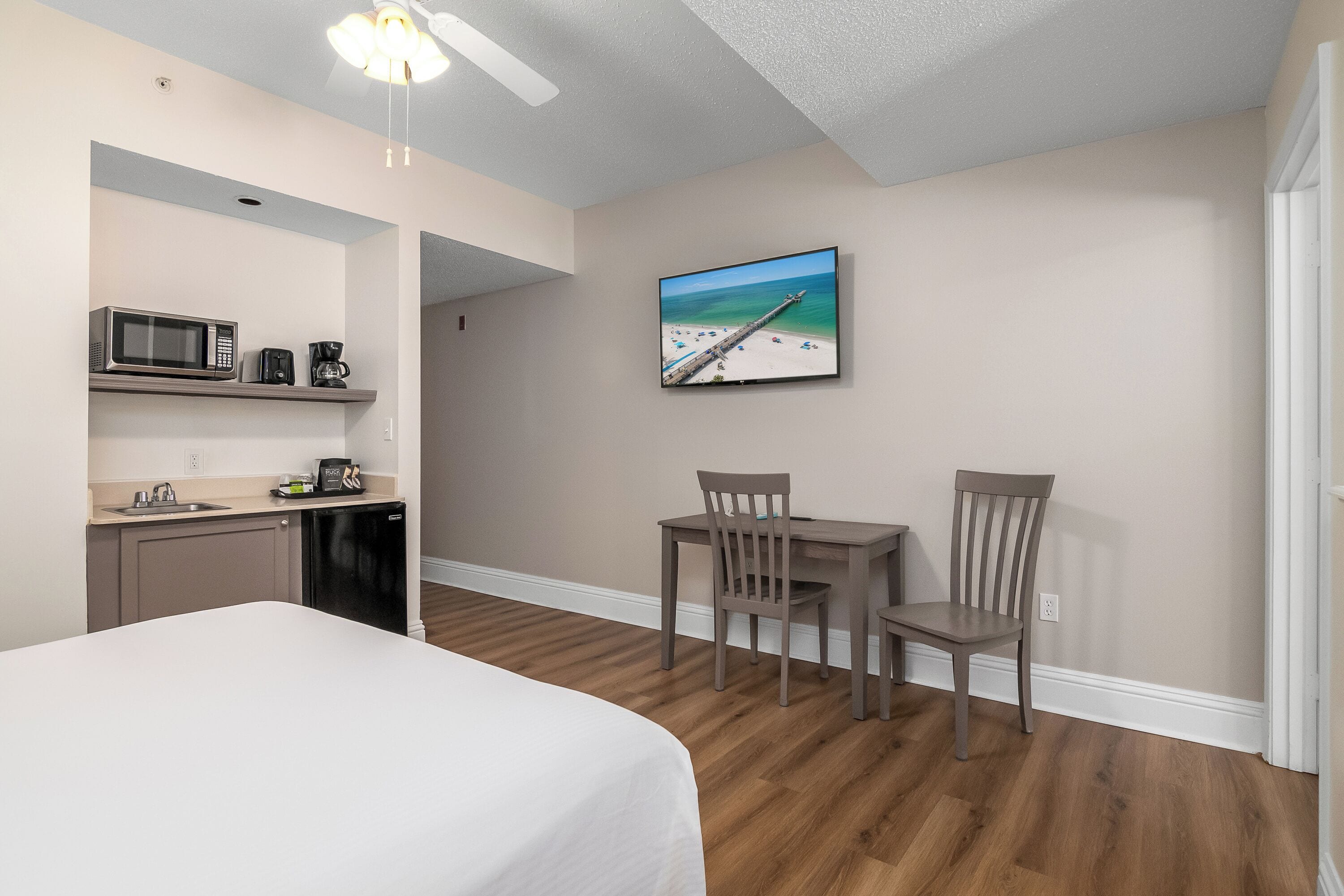 A hotel room with a bed, a kitchenette, a dining area with chairs, and a TV on the wall.