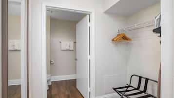 Una habitación con piso de madera, paredes blancas, un banco, un perchero y una puerta que da a un baño con inodoro y toalla.