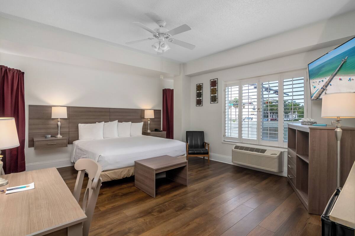 A hotel room with a bed, desk, chair, and a large window with blinds.