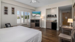 A hotel room with a bed, desk, kitchenette, and a TV displaying a beach scene. - Naples Park Central Hotel (Naples)