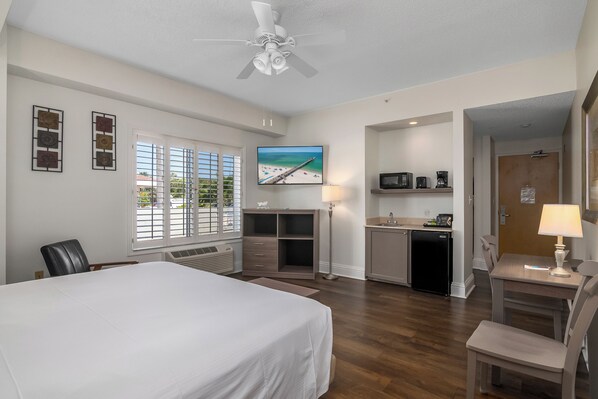 A hotel room with a bed, desk, kitchenette, and a TV displaying a beach scene.