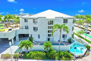 A white multi-story building with a swimming pool, palm trees, and a parking area. - Naples Park Central Hotel (Naples)