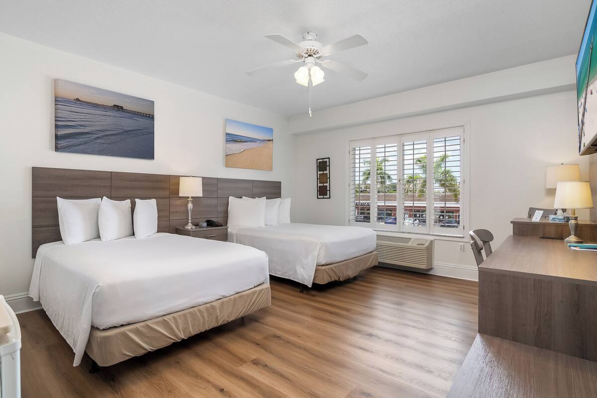 A hotel room with two beds, a desk, a ceiling fan, and large windows with a view of palm trees.