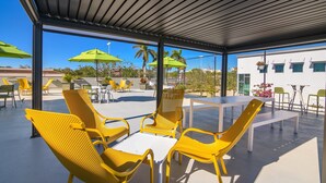 An outdoor patio with yellow chairs and tables, green umbrellas, and a view of buildings and palm trees. - Naples Park Central Hotel (Naples)