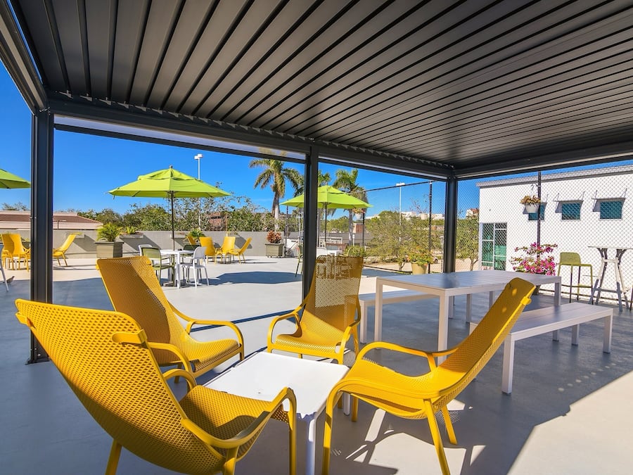 An outdoor patio with yellow chairs and tables, green umbrellas, and a view of buildings and palm trees.