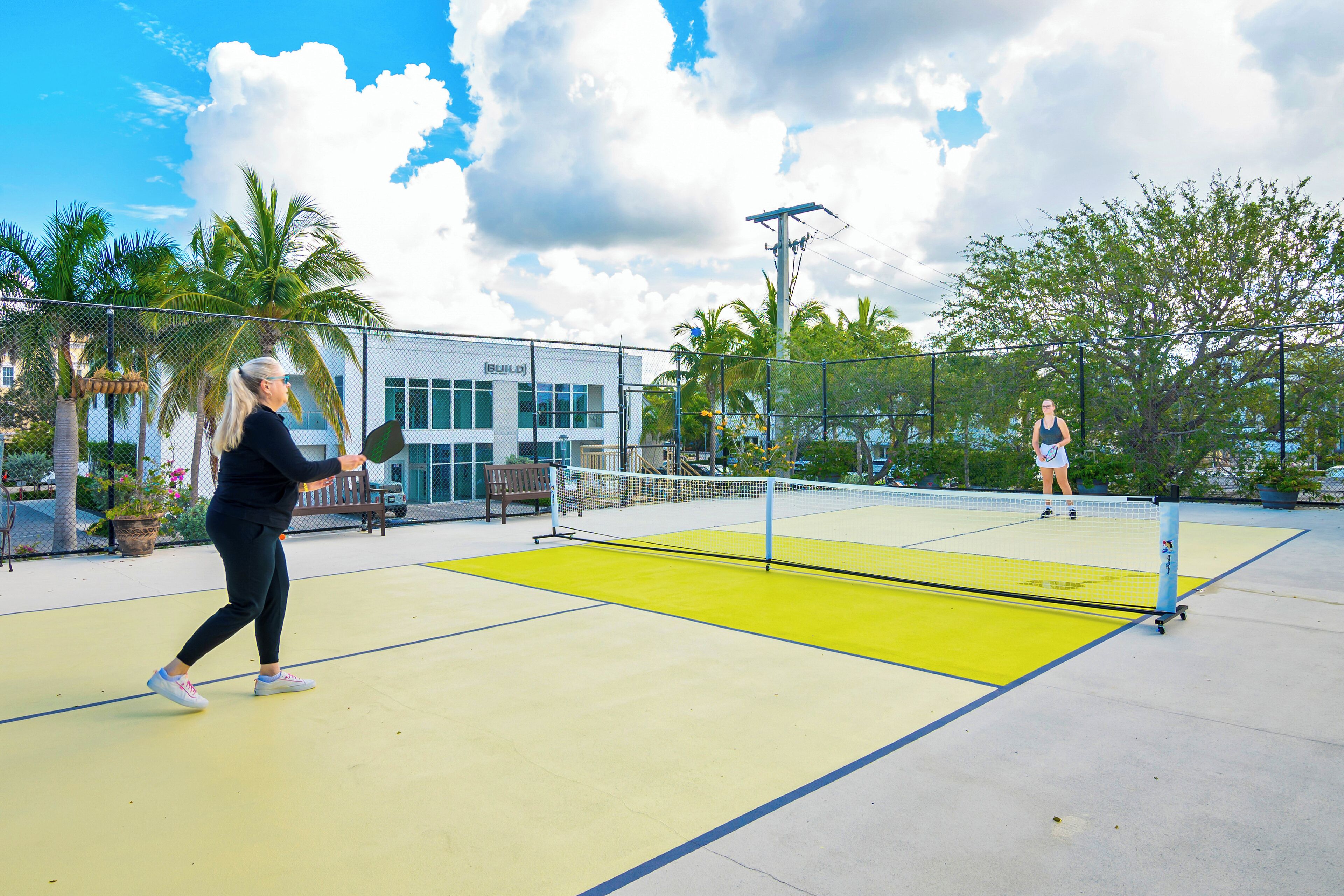 An outdoor tennis court with a net, palm trees, and a building in the background.