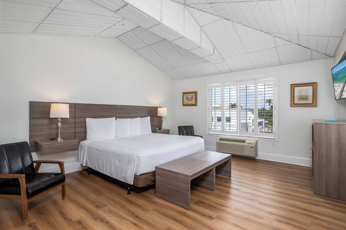 A modern bedroom with a large bed, wooden floor, a bench, a desk, and a TV.