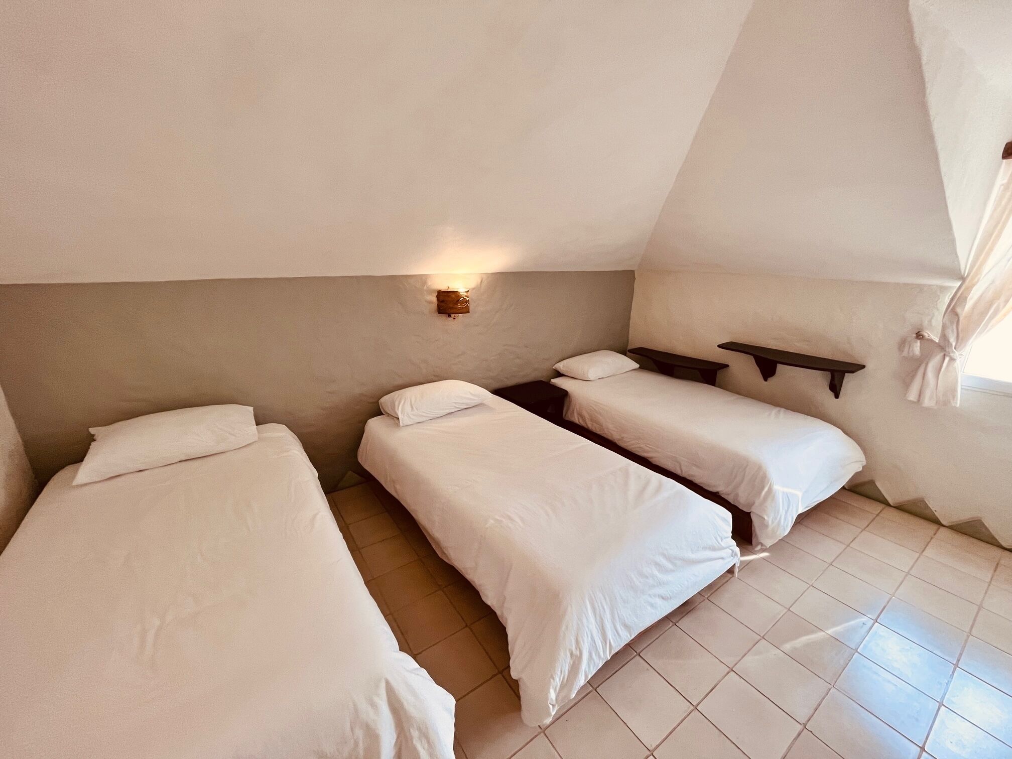 A bedroom with two single beds, a bedside table, a wall-mounted shelf, and a window with curtains.