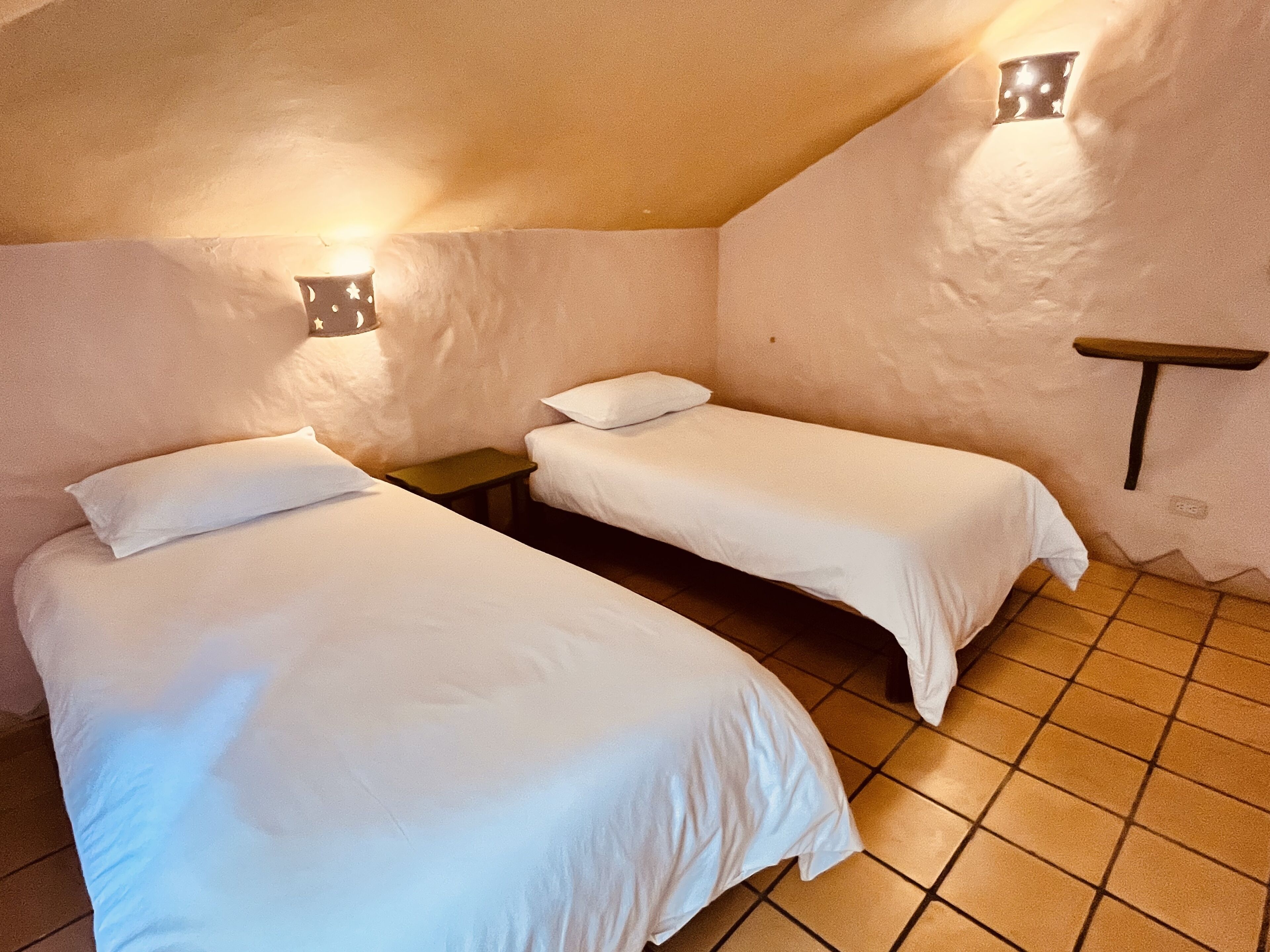 Ein kleines, gemütliches Zimmer mit zwei Betten, einem Nachttisch und einem an der Wand befestigten Regal.